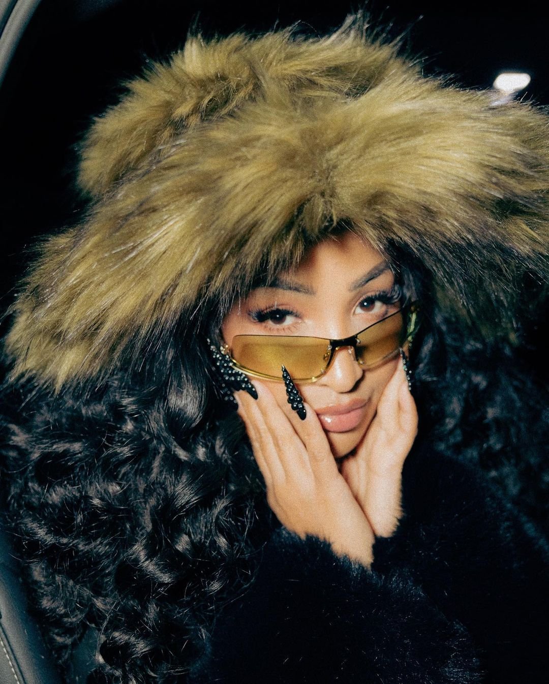Portrait of a woman in a furry hat and yellow sunglasses, holding her hands up to her face in a studio with a dark background.