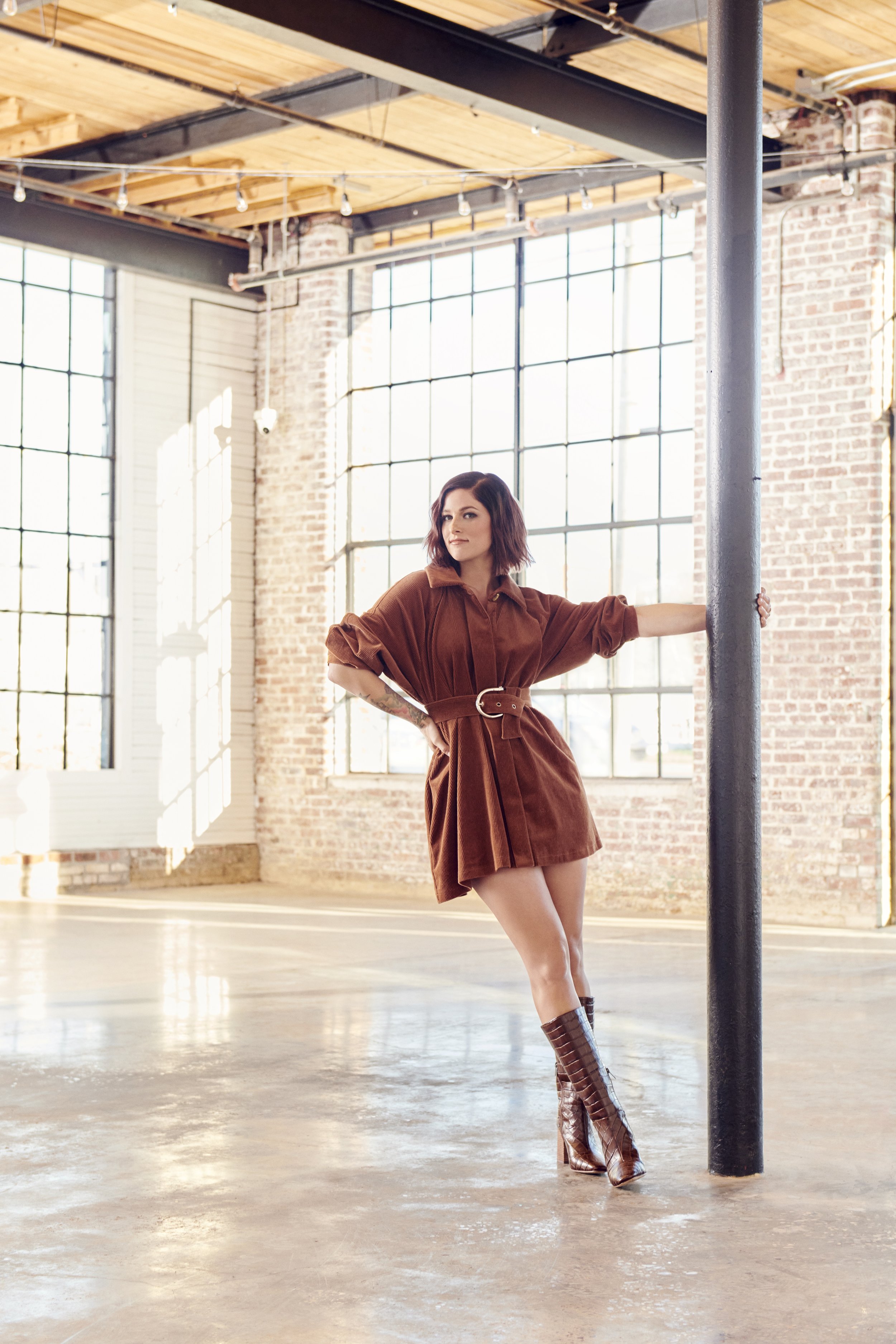 A stylish woman poses in a brown corduroy dress and matching boots, leaning against a pillar in a sunlit industrial loft interior.