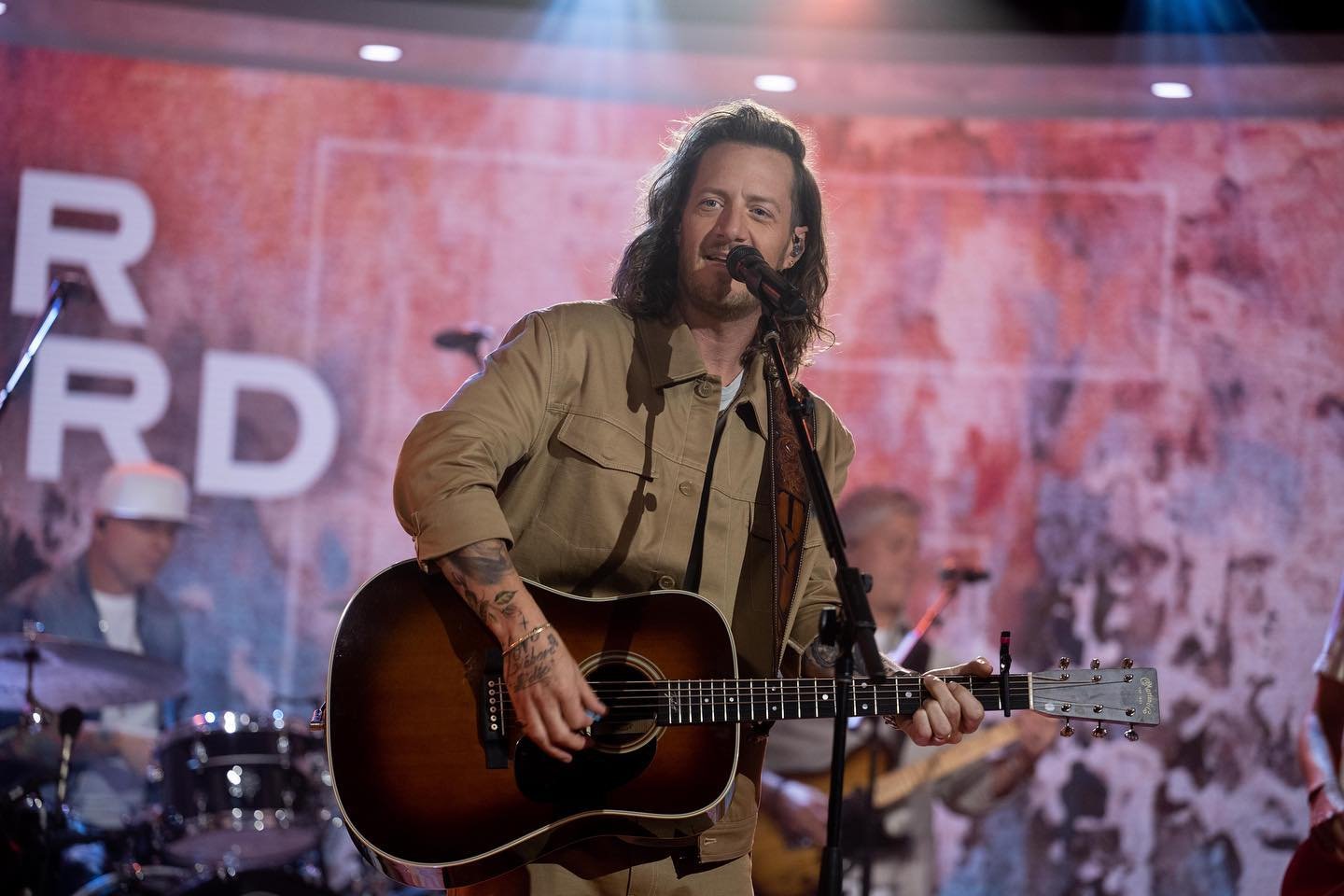 A solo male music artist performs on stage with an acoustic guitar, backed by a band during an indoor concert with stage lighting.