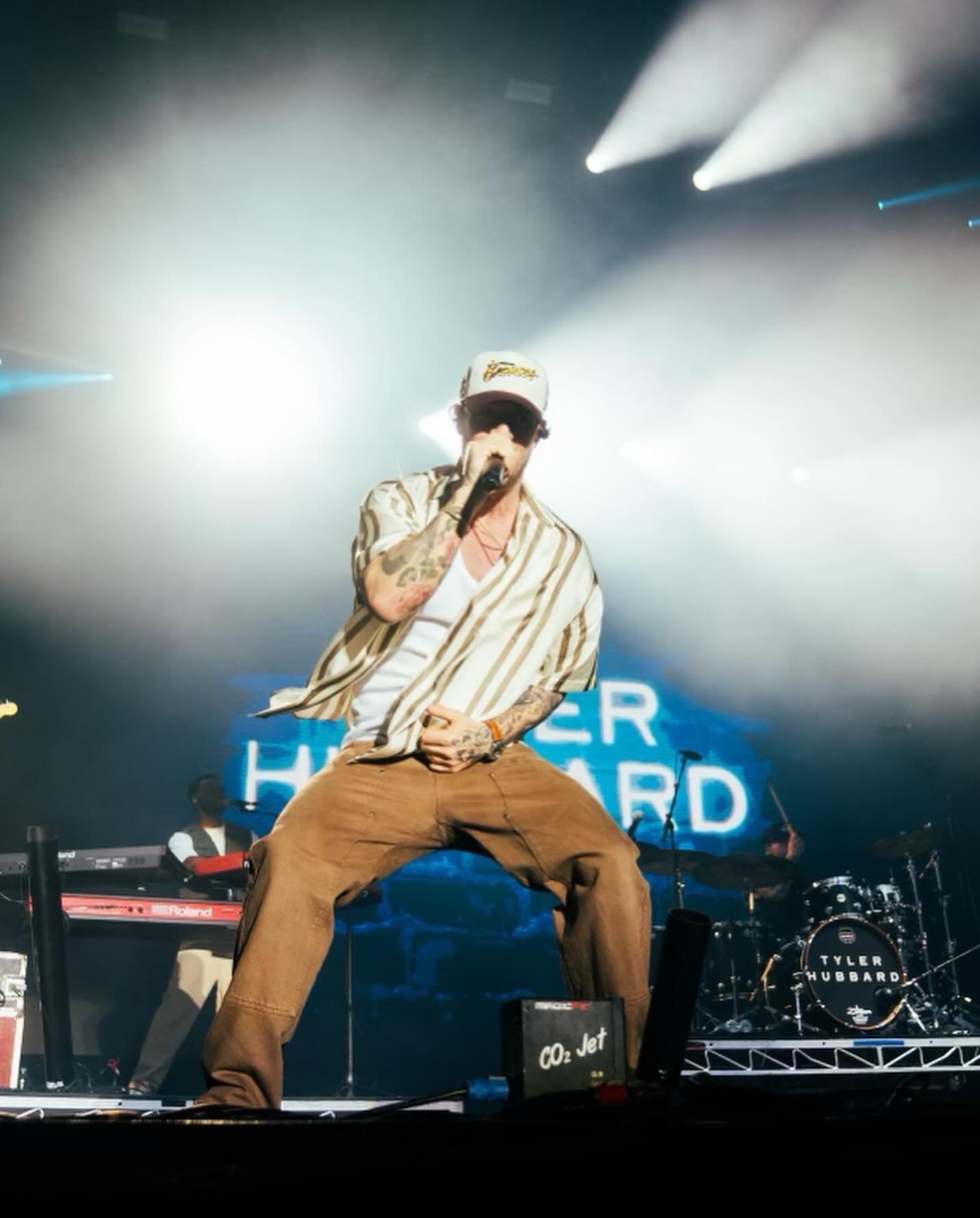 Tyler Hubbard performing live on stage at a concert. He is singing into a microphone while wearing a hat and striped shirt.