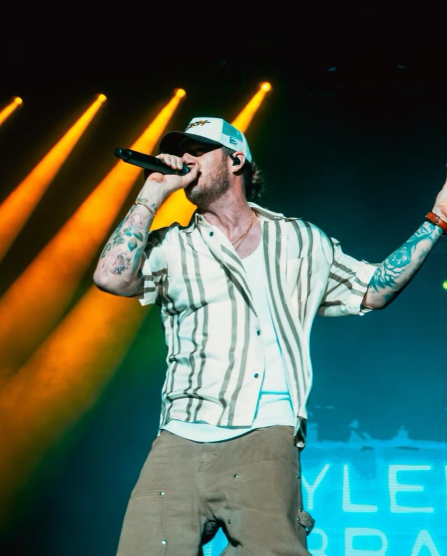 An adult male singer with tattoos and a baseball cap performs on stage with lights. He wears a striped shirt and khaki pants.