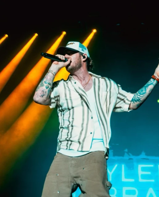 An adult male singer with tattoos and a baseball cap performs on stage with lights. He wears a striped shirt and khaki pants.