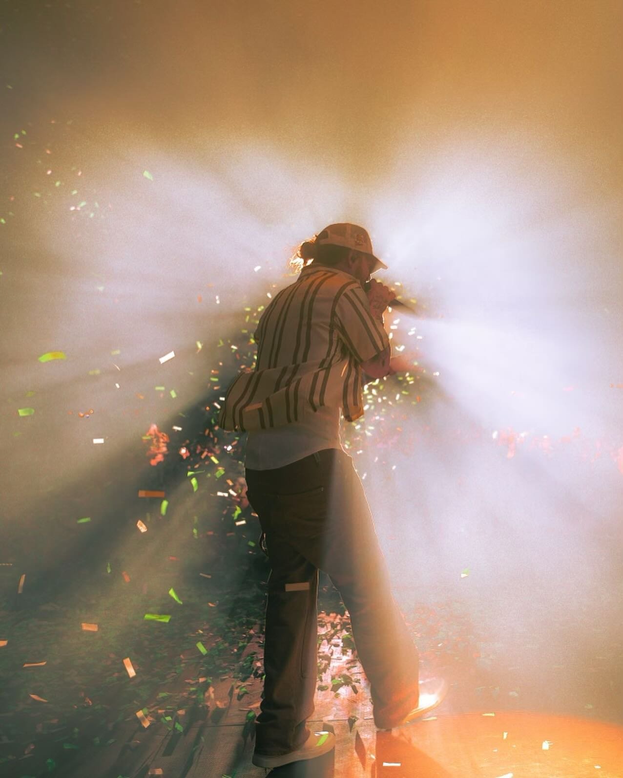 Male musician performs on stage with confetti and dynamic lighting, wearing a hat, striped shirt, and light-colored jeans.