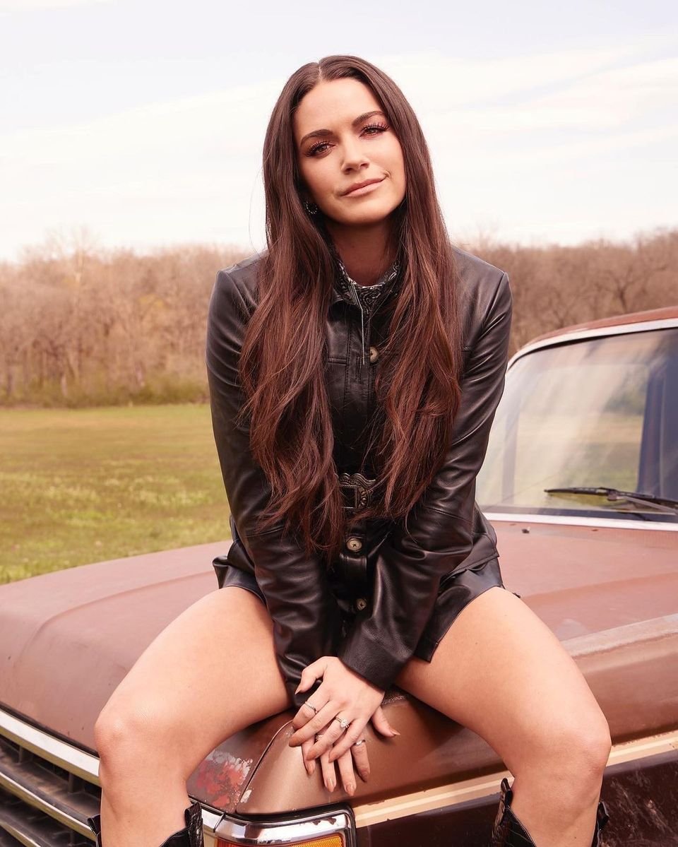 An adult female with long brown hair, wearing a black jacket, is sitting on the hood of a brown pickup truck in a grassy field.