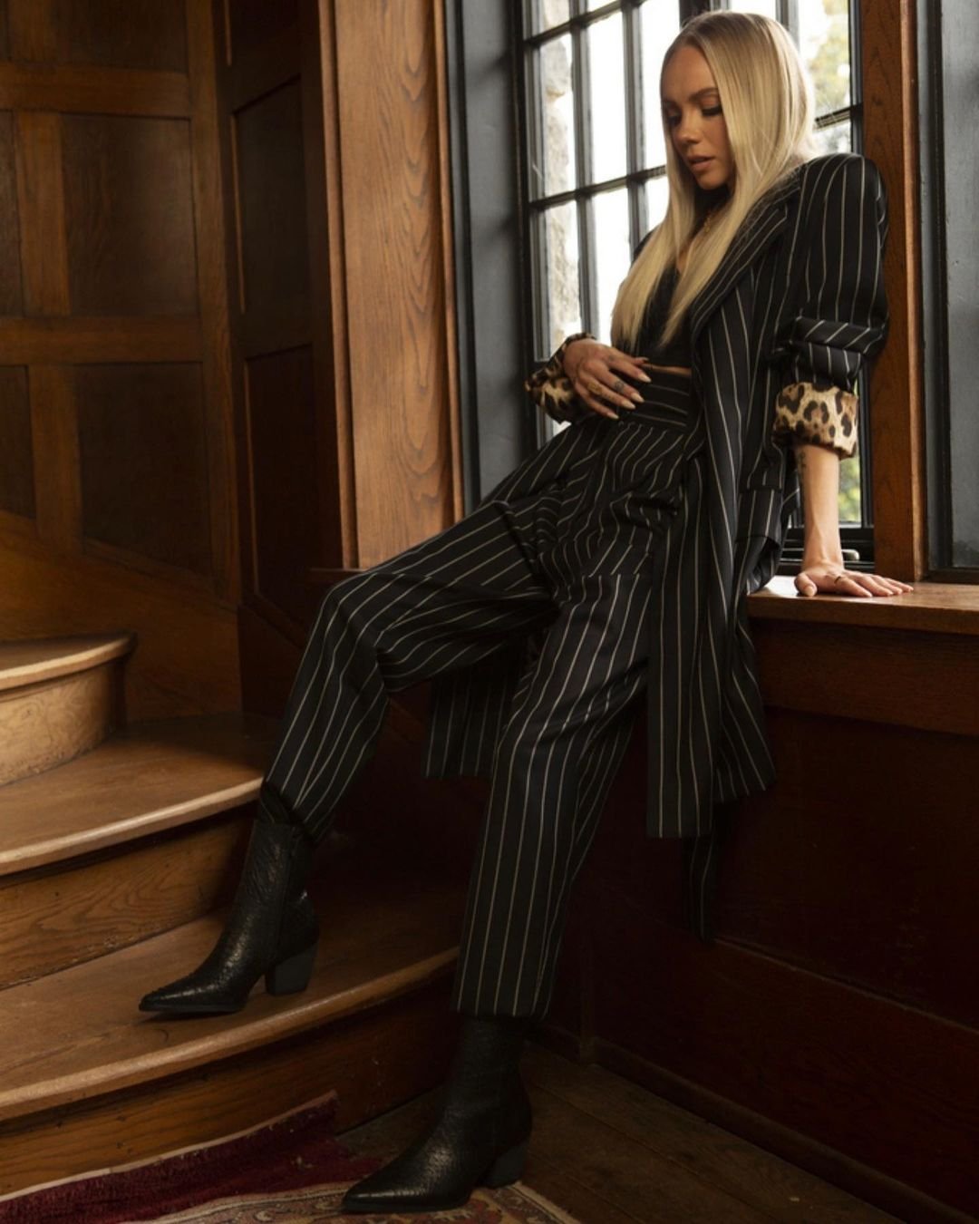 Blonde woman in a pinstripe suit poses near a window, showcasing high-fashion style inside a wooden staircase environment.