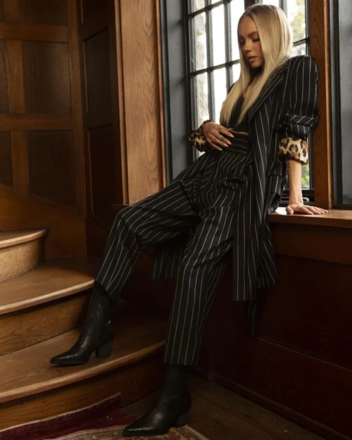Blonde woman in a pinstripe suit poses near a window, showcasing high-fashion style inside a wooden staircase environment.