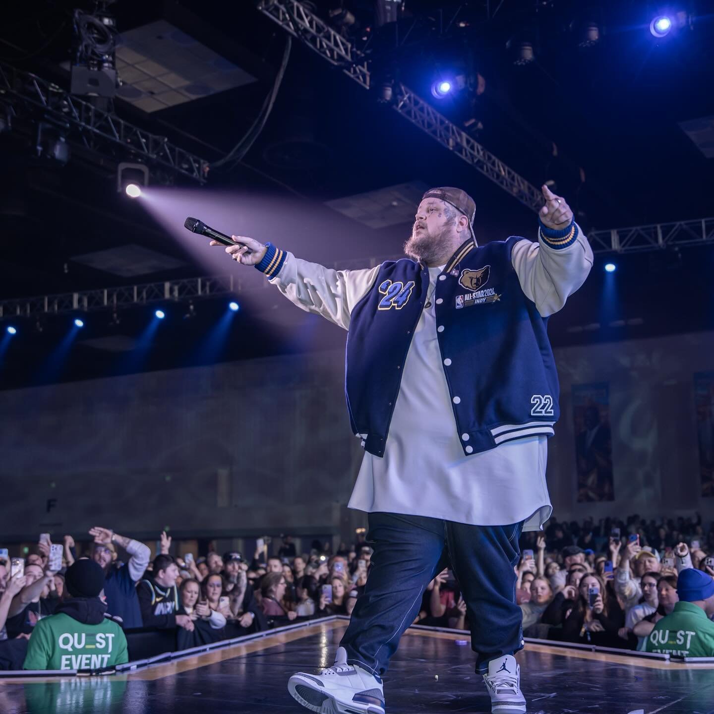 A male musician wearing a blue and white jacket is performing live on stage in front of a large crowd, holding a microphone.