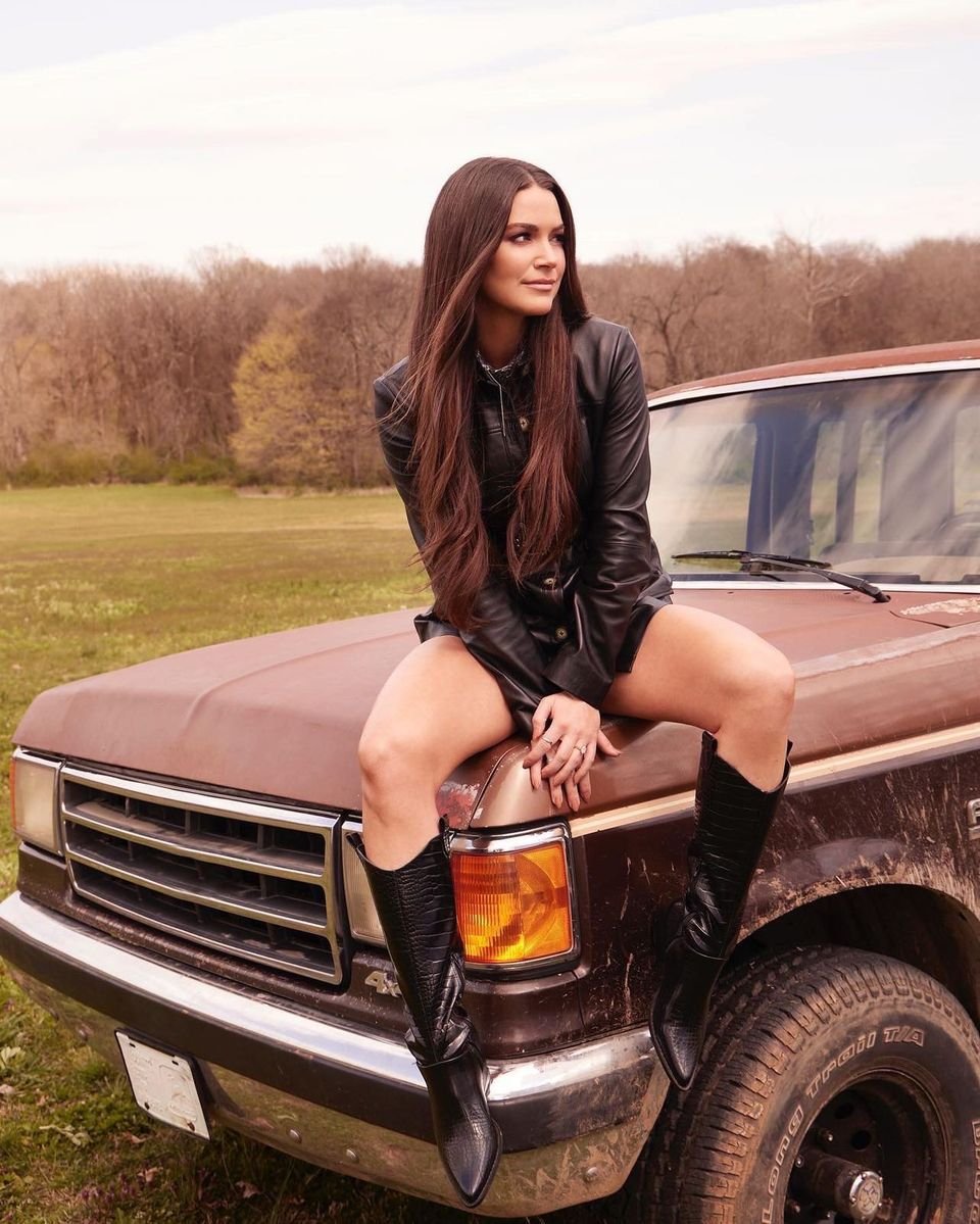 An adult woman poses on the hood of a brown truck outdoors in a field, wearing a black leather jacket and black boots.