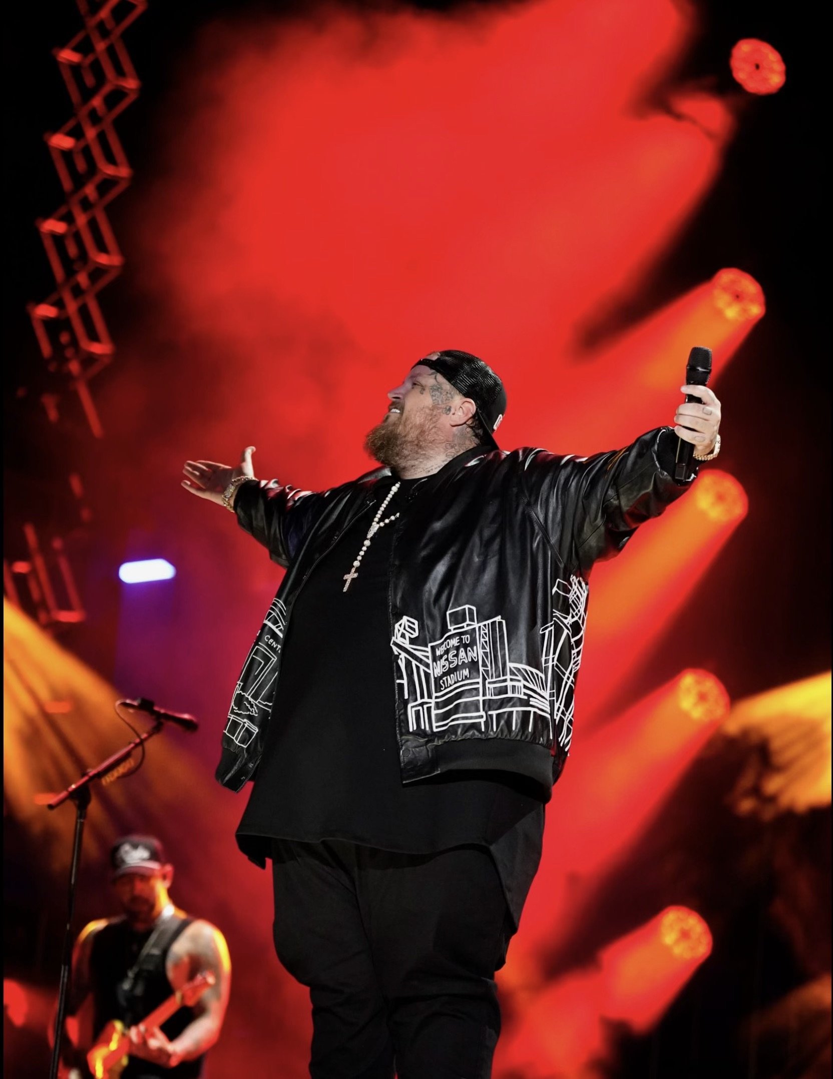 Bearded singer performs live on stage with a microphone, wearing a black jacket with white building outlines, with red stage lighting.