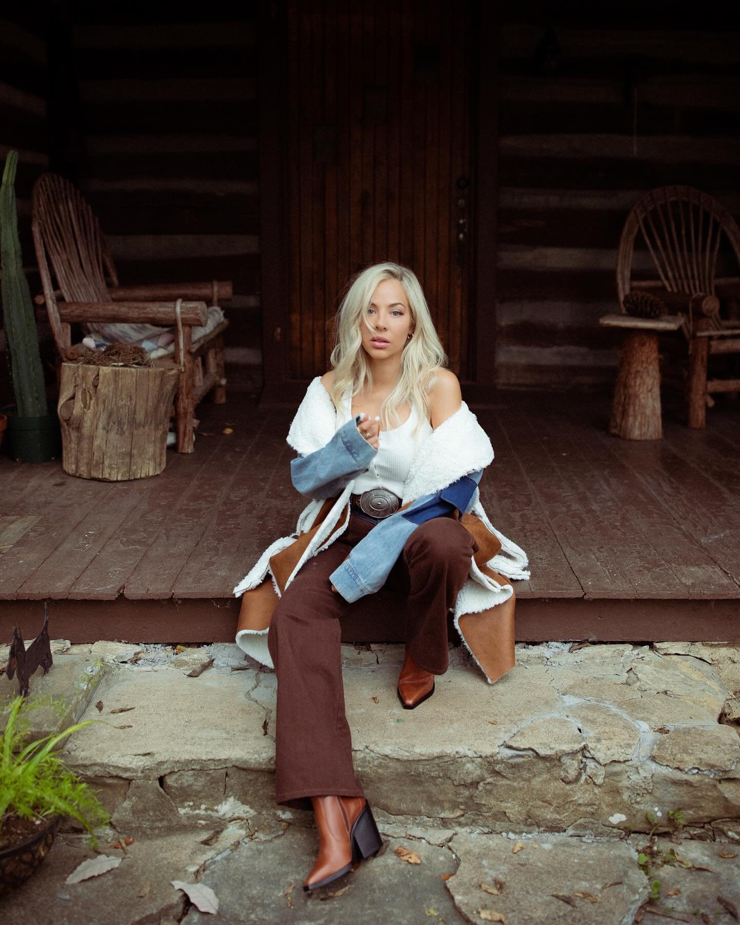 Blonde woman in a white top and brown pants sits on a stone step of a log cabin, wearing boots and a denim coat with shearling.