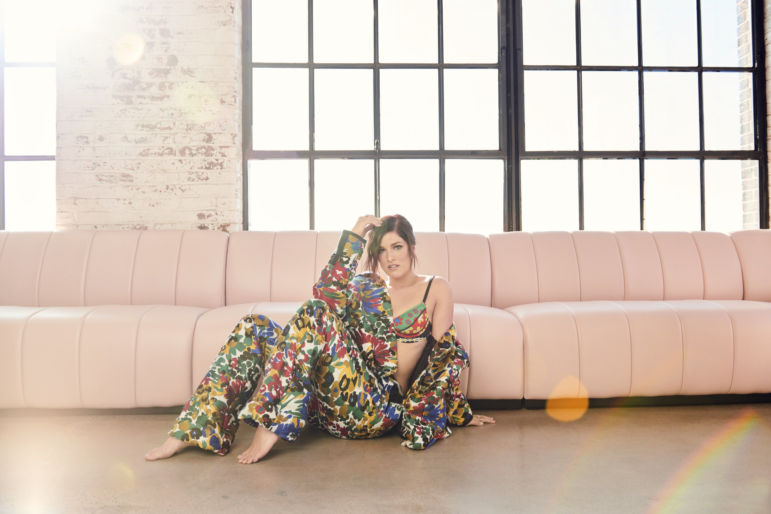 Brunette woman in floral pants and bra top sitting on floor next to pink sofa in bright, modern room with large windows and exposed brick.