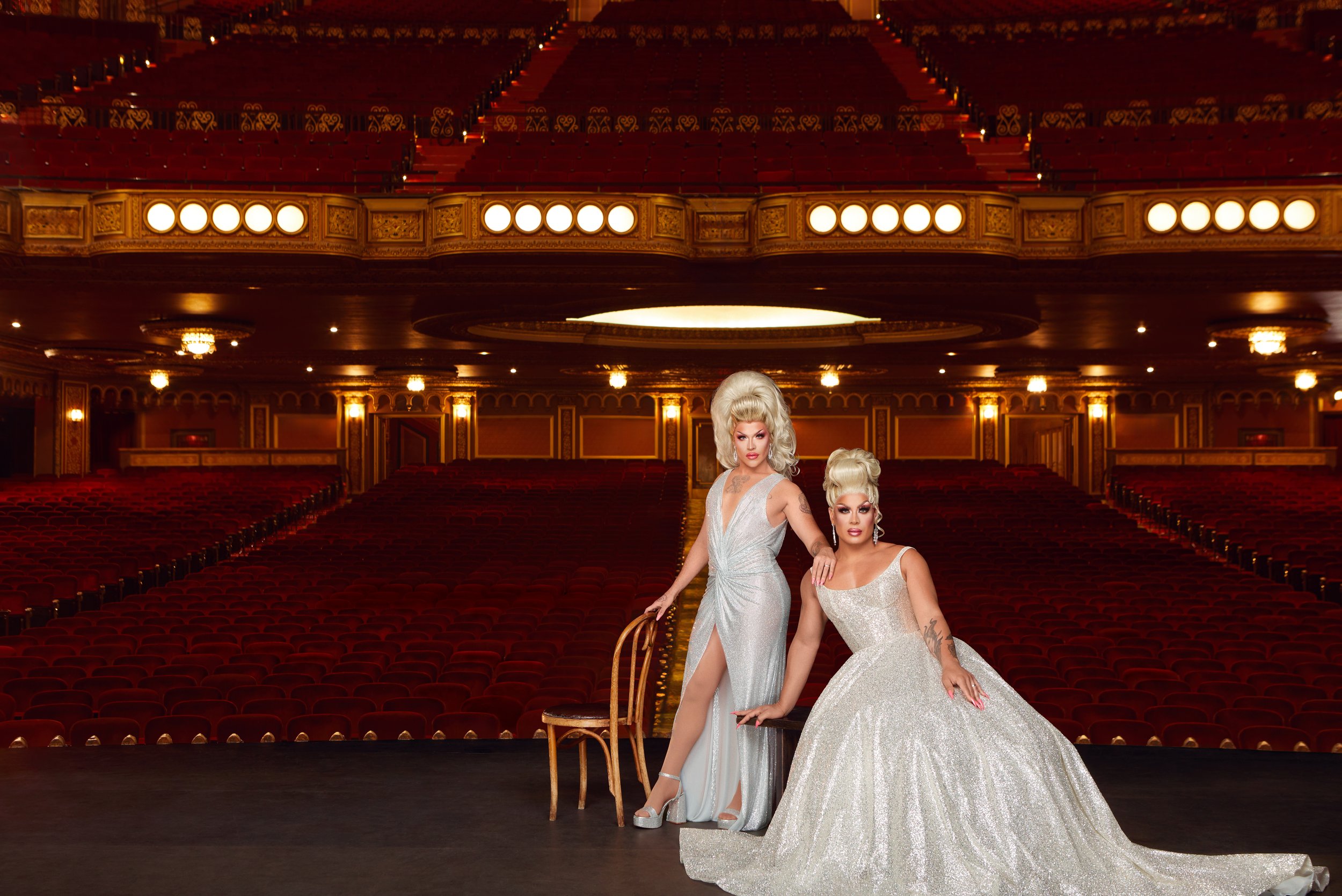 Two drag queens in elegant gowns pose on a theater stage with red velvet seating. Ornate gold details enhance the grand interior.