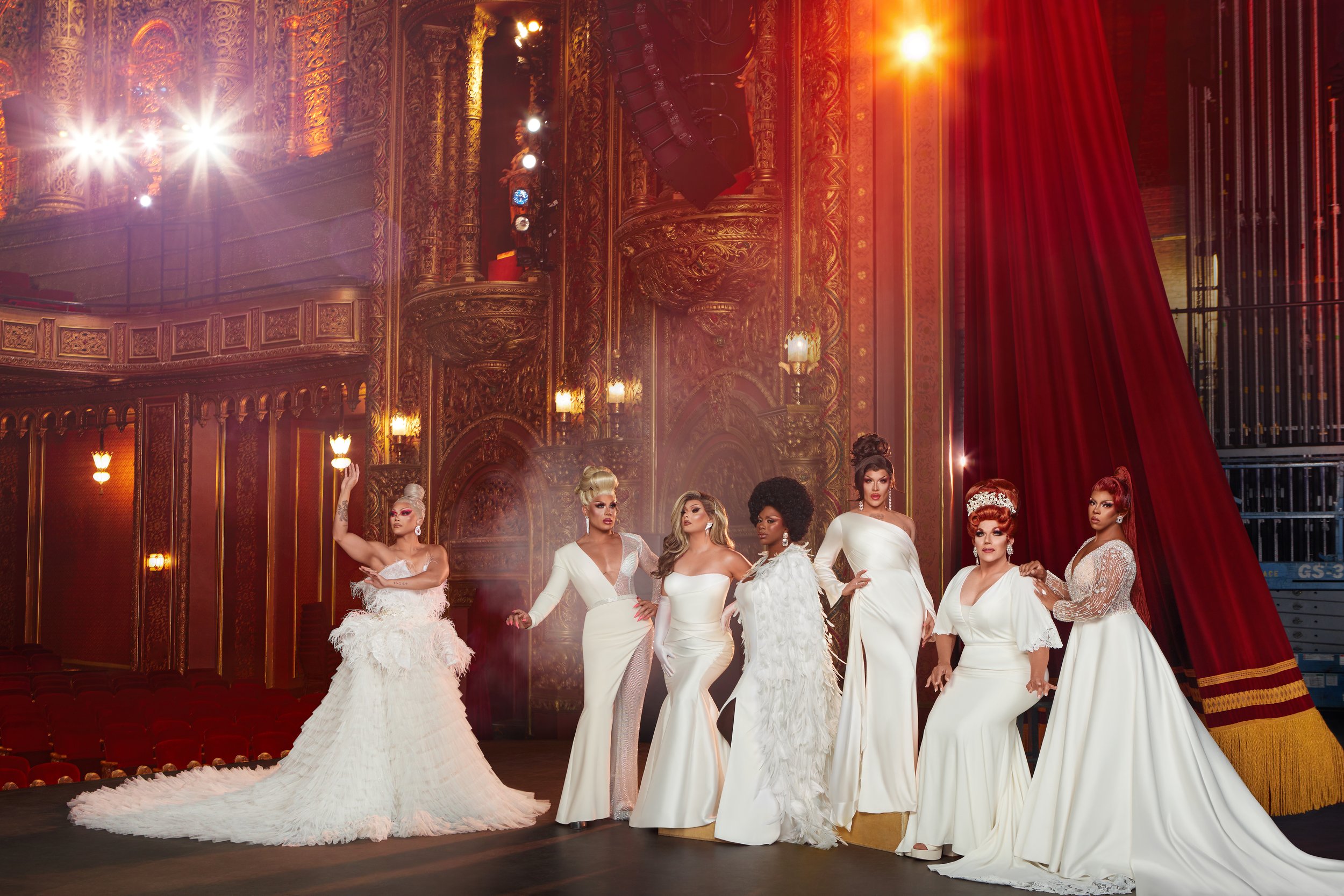 Seven drag queens wearing elegant white gowns pose on stage with ornate gold details and red curtains, illuminated by bright stage lights.