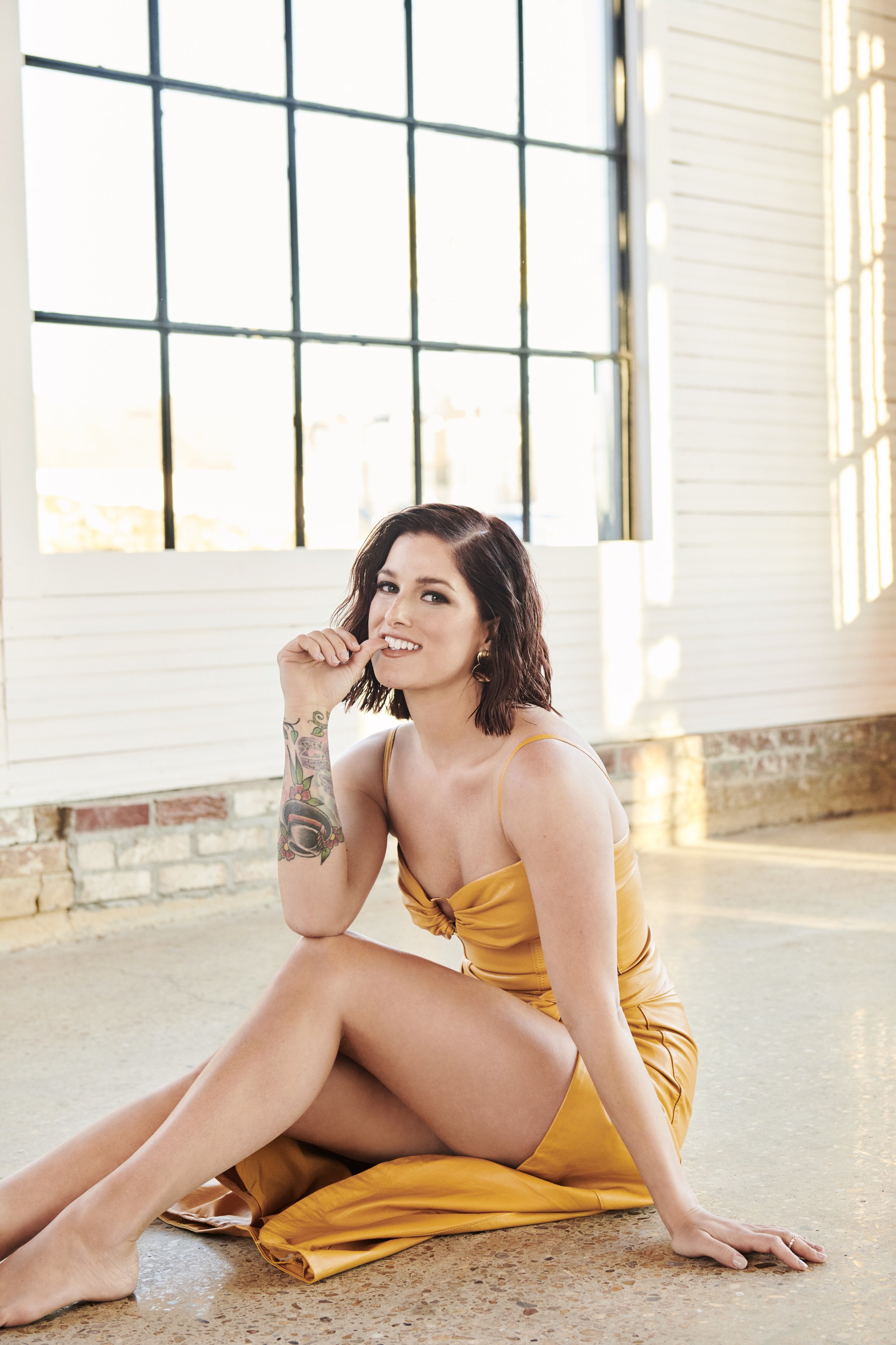 Woman in gold dress sits in front of a large window in a bright interior space, smiling and looking at the camera, with natural light.