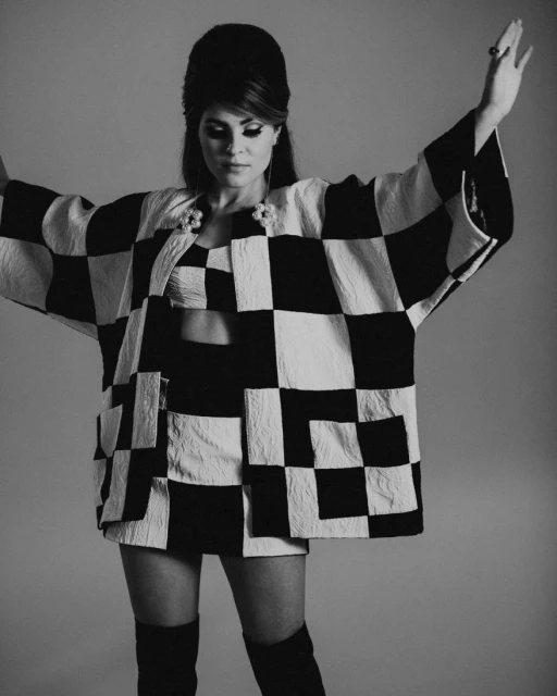 A woman with a bouffant hairstyle poses in a black and white checkered jacket, crop top, mini skirt, and over-the-knee boots against a gray backdrop.