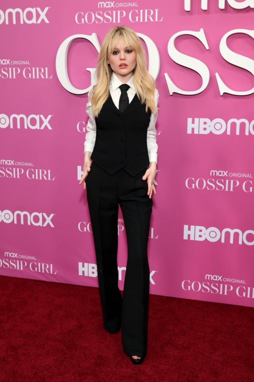 Blonde woman in black vest and pants on a pink Gossip Girl branded carpet poses confidently, showcasing fashion and event attire.