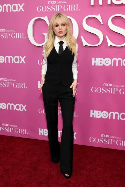 Blonde woman in black vest and pants on a pink Gossip Girl branded carpet poses confidently, showcasing fashion and event attire.