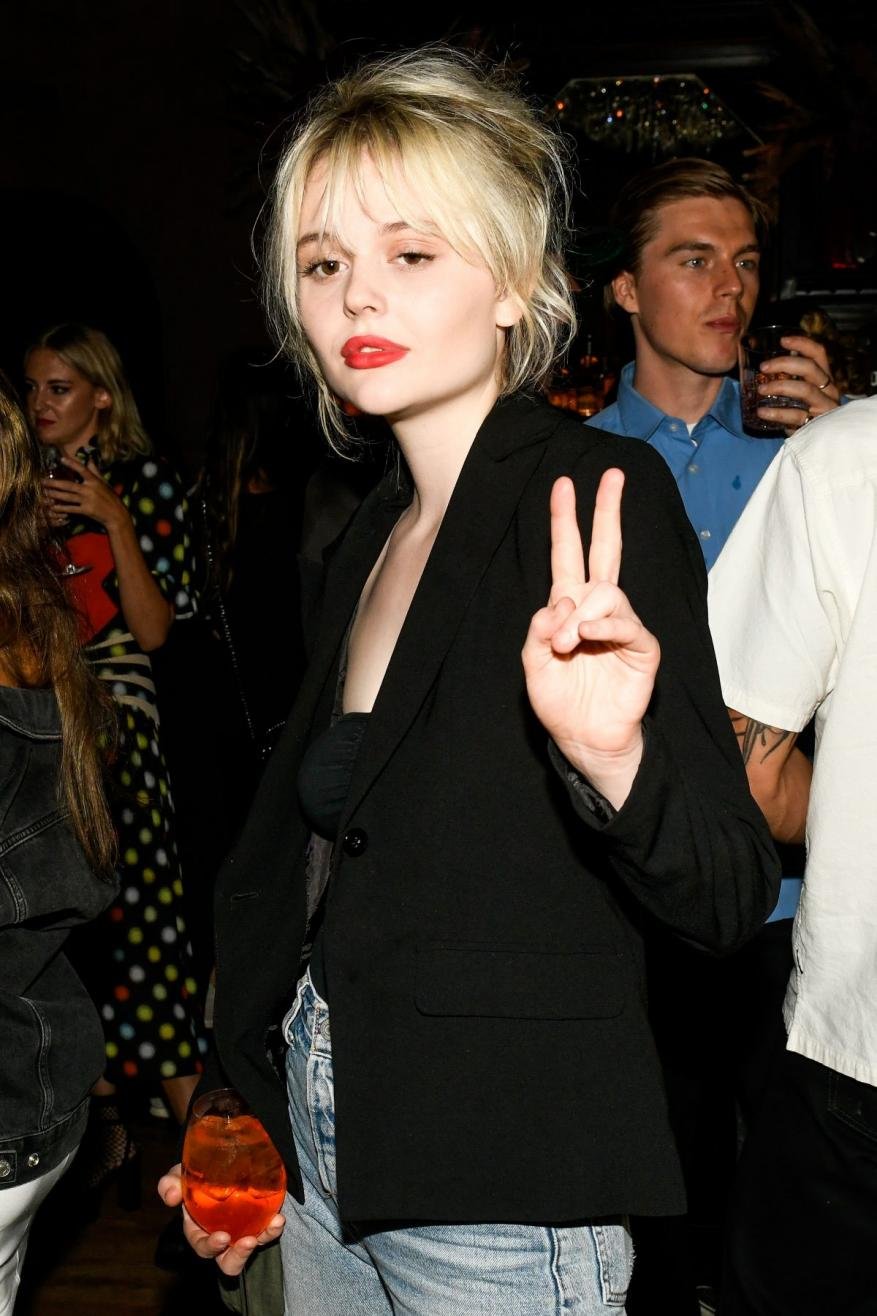 Woman with blonde hair and red lipstick holds a cocktail glass and makes a peace sign in a dimly lit indoor environment.