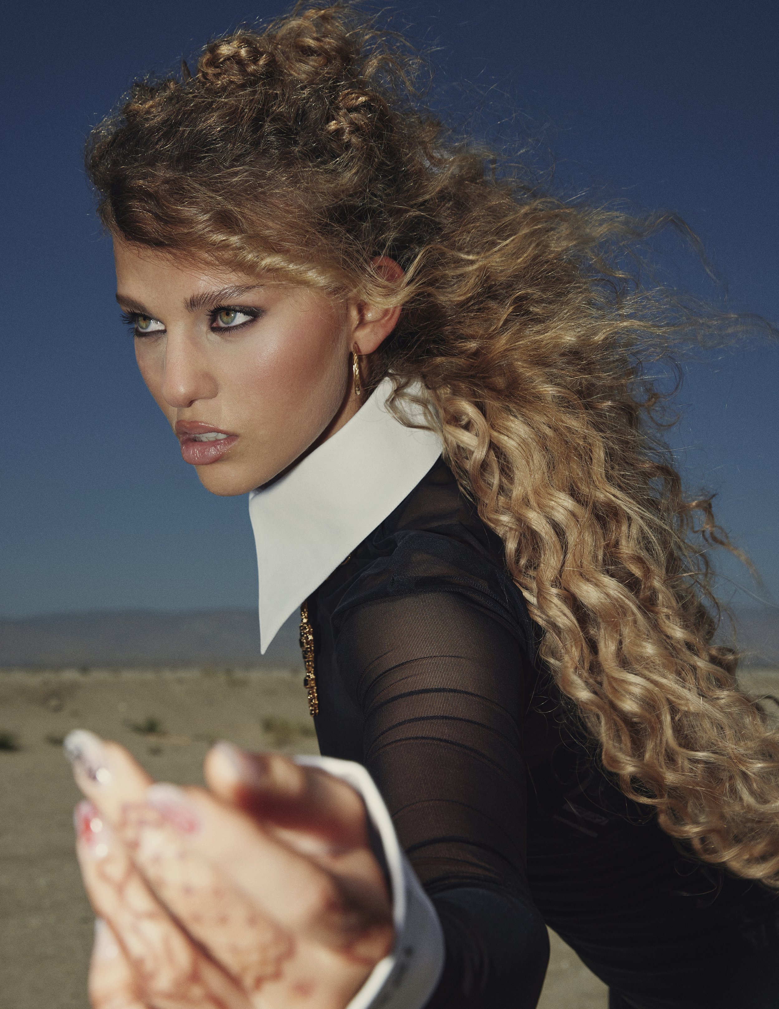 Woman with curly blonde hair wearing a black sheer top and white collar standing in a desert with a blue sky background.