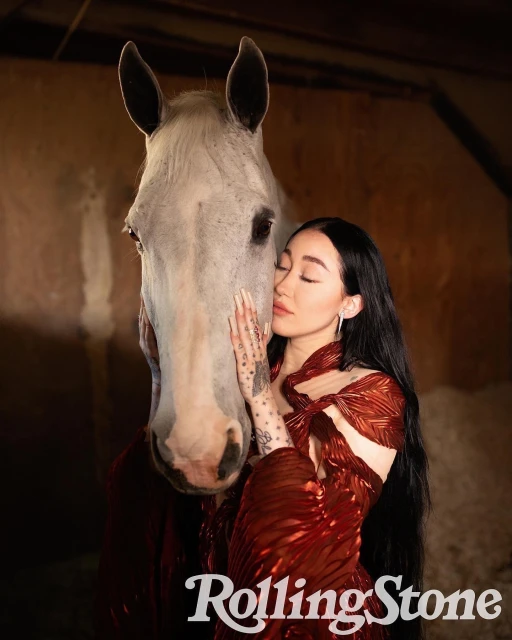 Portrait of a woman with long black hair in a red dress embracing a white horse indoors, with a Rolling Stone logo in the corner.