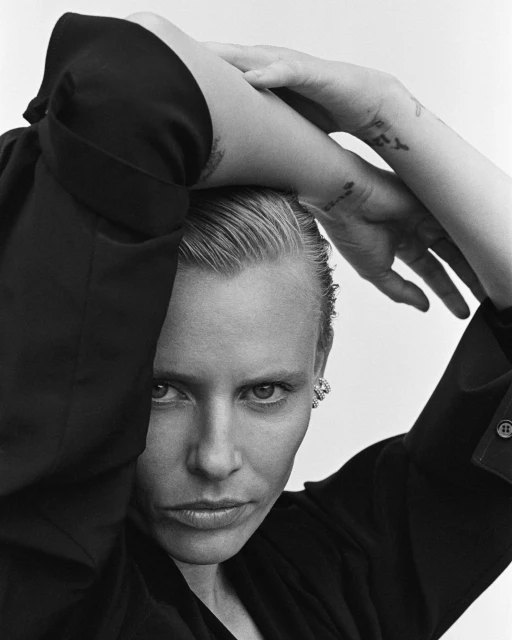 Black-and-white portrait of a woman with short blonde hair, dark shirt, and tattoos posing indoors in front of a white background.