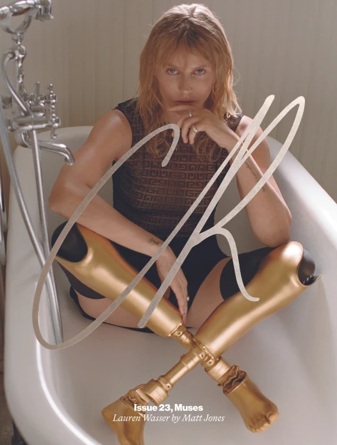 Blonde woman poses in a white bathtub with crossed golden prosthetic legs, wearing a dark top in an indoor setting with natural light.