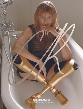 Blonde woman poses in a white bathtub with crossed golden prosthetic legs, wearing a dark top in an indoor setting with natural light.