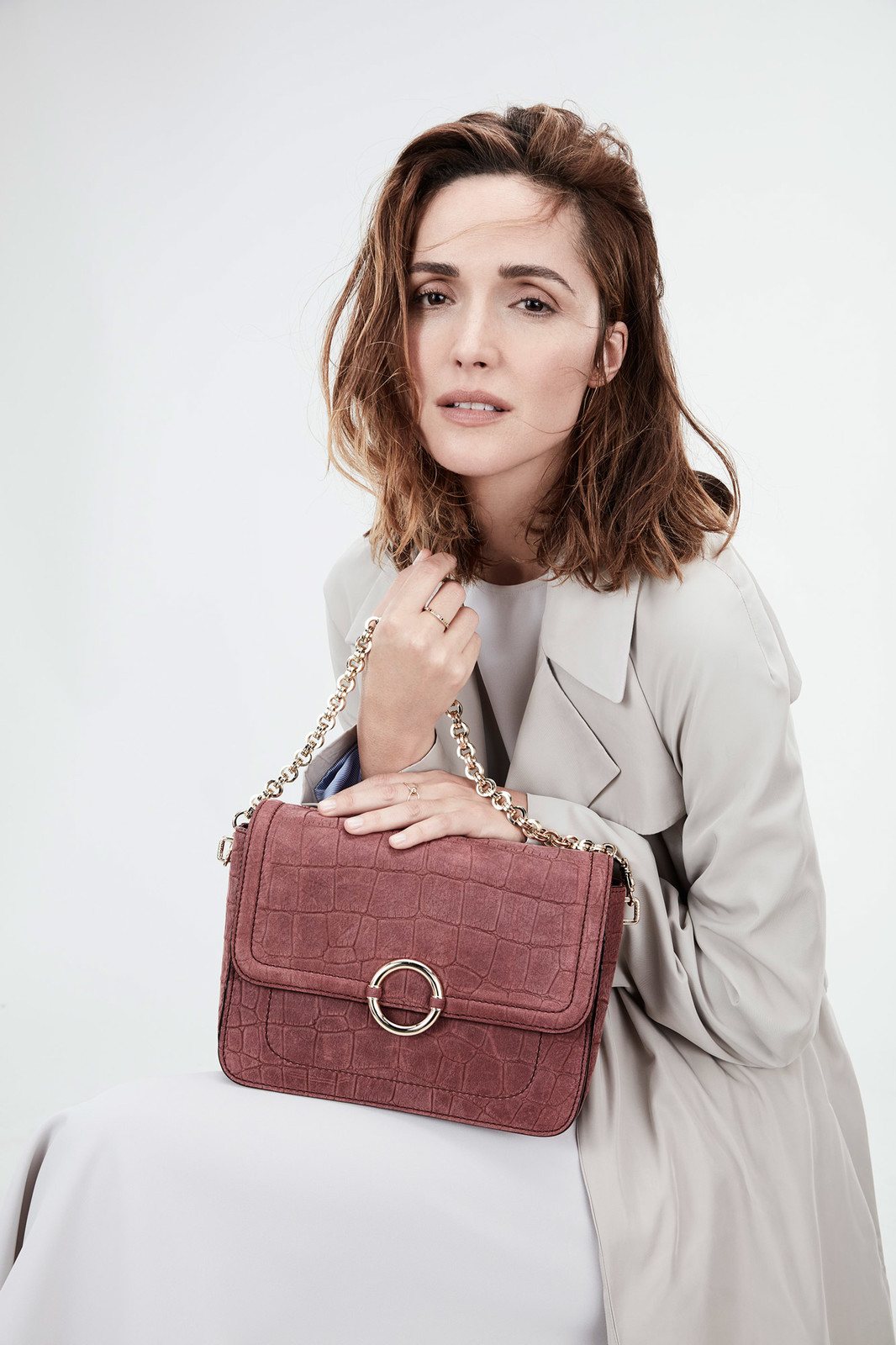 Elegant woman poses with a stylish red leather purse with gold chain. She wears a beige trench coat against a clean, bright background.