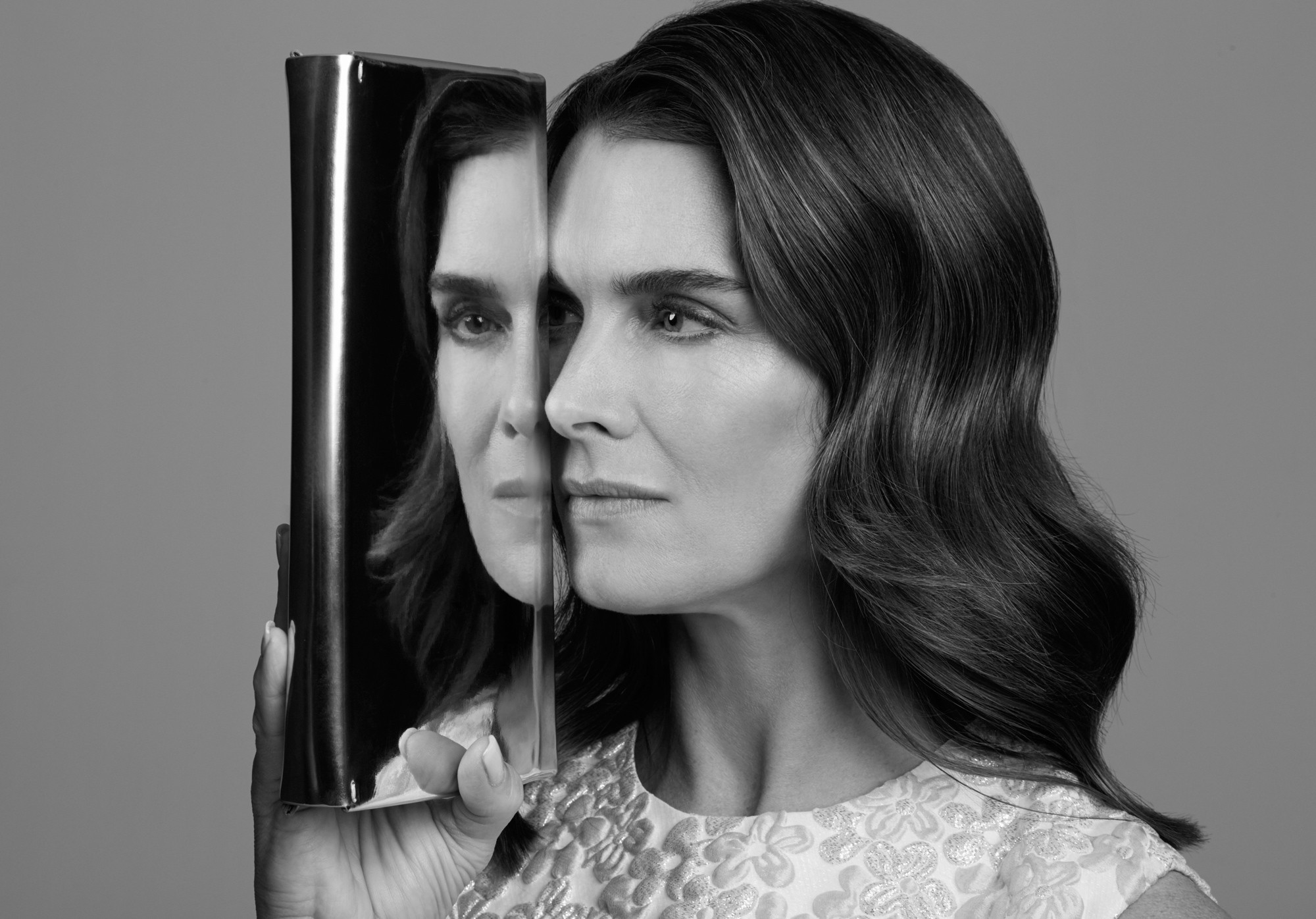 Monochrome portrait of a woman holding a mirror reflecting half her face, set against a neutral gray background. Beauty and reflection.