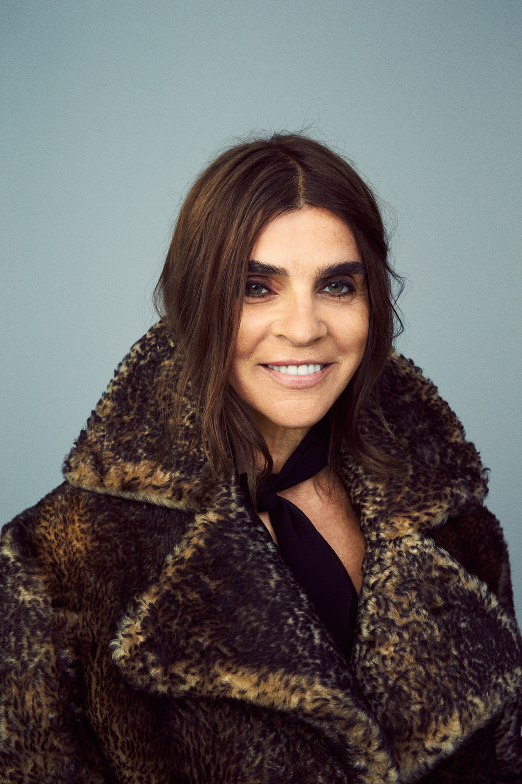 Headshot of an adult woman with brown hair, wearing a patterned fur coat against a grey background, smiling at the camera.