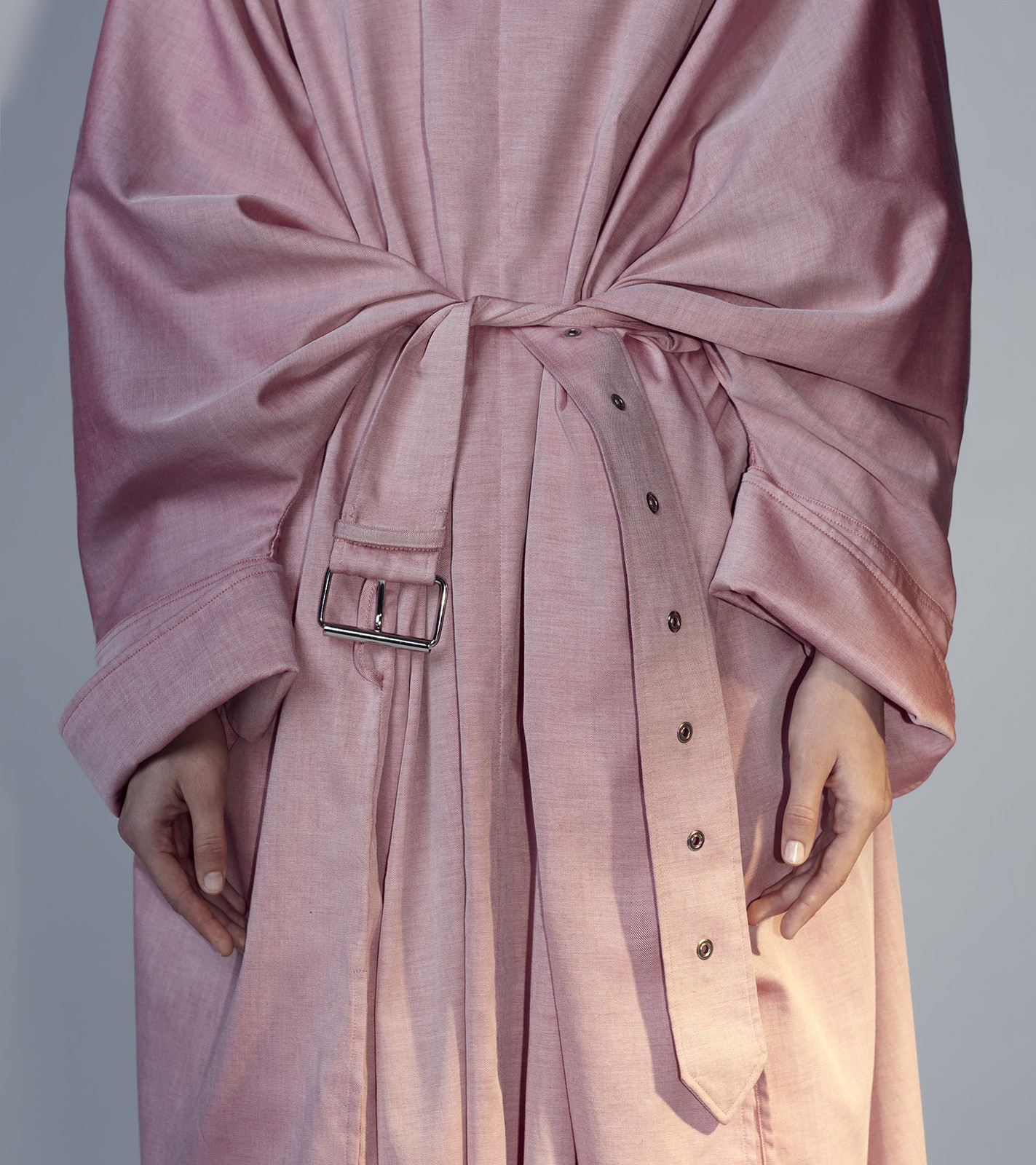Close-up of a model wearing a pink trench coat tied at the waist, showcasing the fabric texture, belt, and design details.