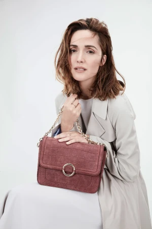 Rose Byrne wearing a light grey coat holding a textured mauve shoulder bag against a white backdrop