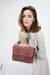 Rose Byrne wearing a light grey coat holding a textured mauve shoulder bag against a white backdrop