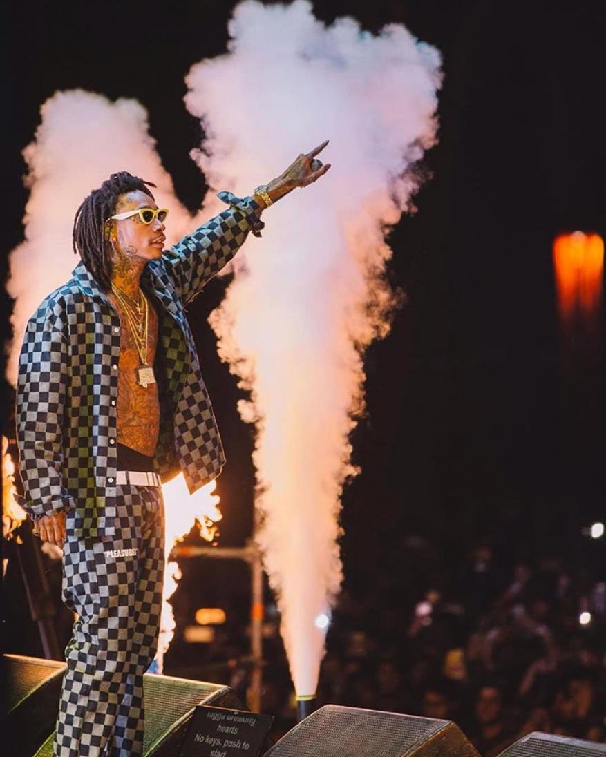 Wiz Khalifa performing on stage in a black-and-white checkered suit, with bright lights and smoke effects behind him during a concert.