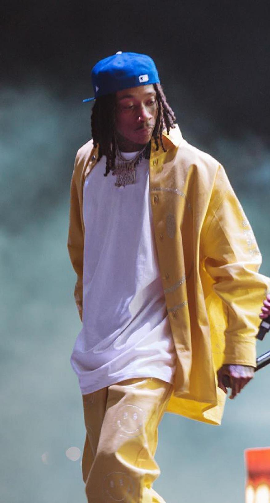 Adult male singer performing on stage in a blue baseball cap, white t-shirt, and yellow jacket and pants with smoky background.