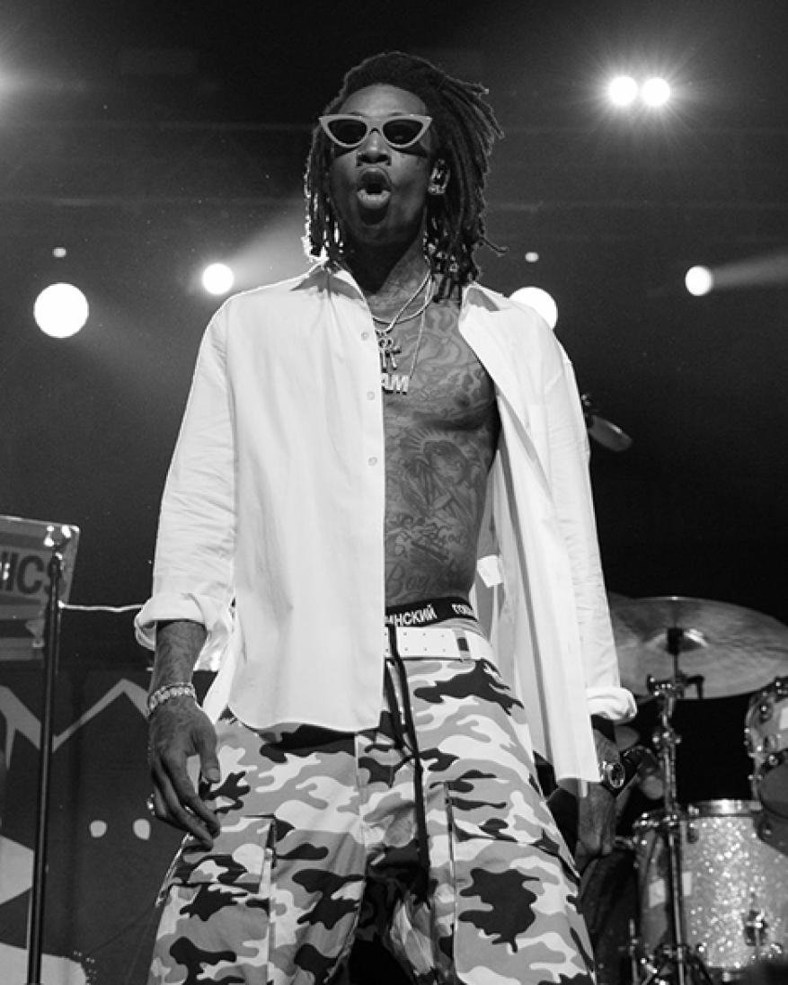 Wiz Khalifa performing on stage, wearing sunglasses, white shirt, and camouflage pants, in a black and white concert photo.
