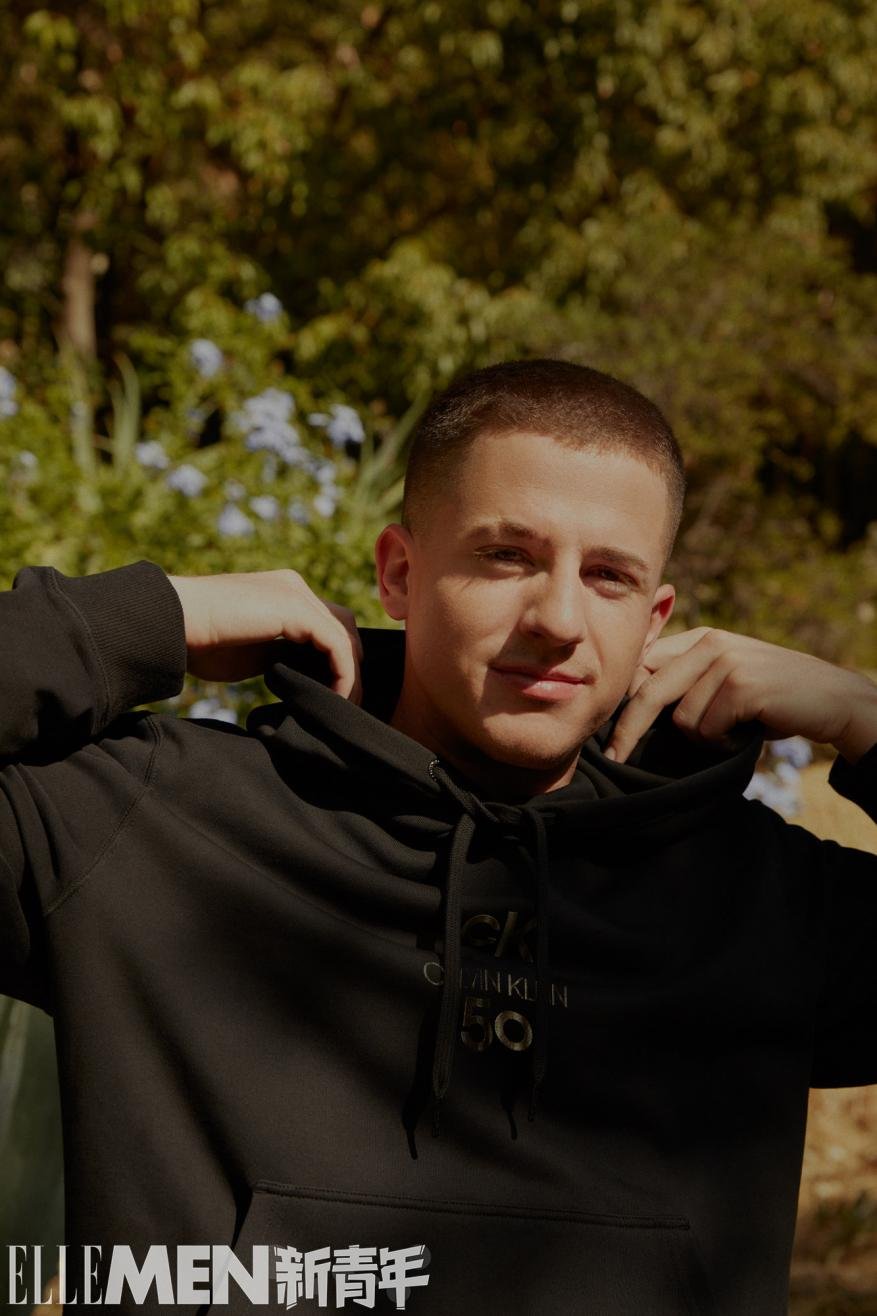 Portrait of a smiling adult male in a black Calvin Klein hooded sweatshirt outdoors with a blurred background of green trees and plants.