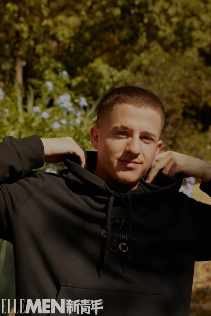 Portrait of a smiling adult male in a black Calvin Klein hooded sweatshirt outdoors with a blurred background of green trees and plants.