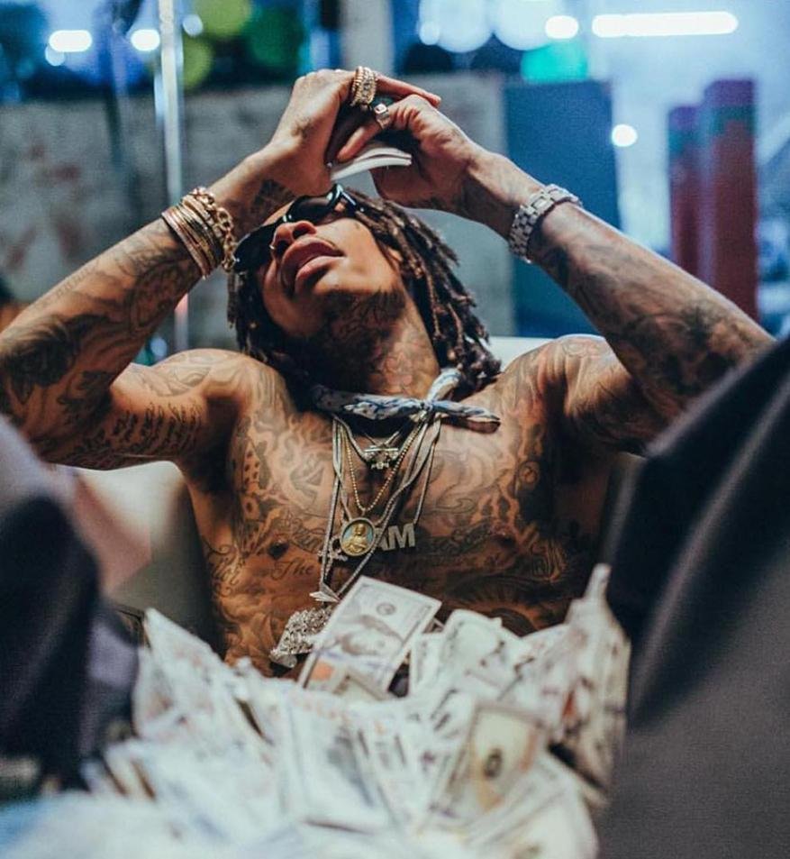 Shirtless man with dreadlocks wearing jewelry, holding money in an indoor setting filled with money and wearing sunglasses.