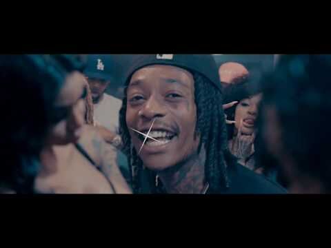 Close-up of Wiz Khalifa, an adult male wearing a black cap and necklace. He is surrounded by people with various tattoos and jewelry.