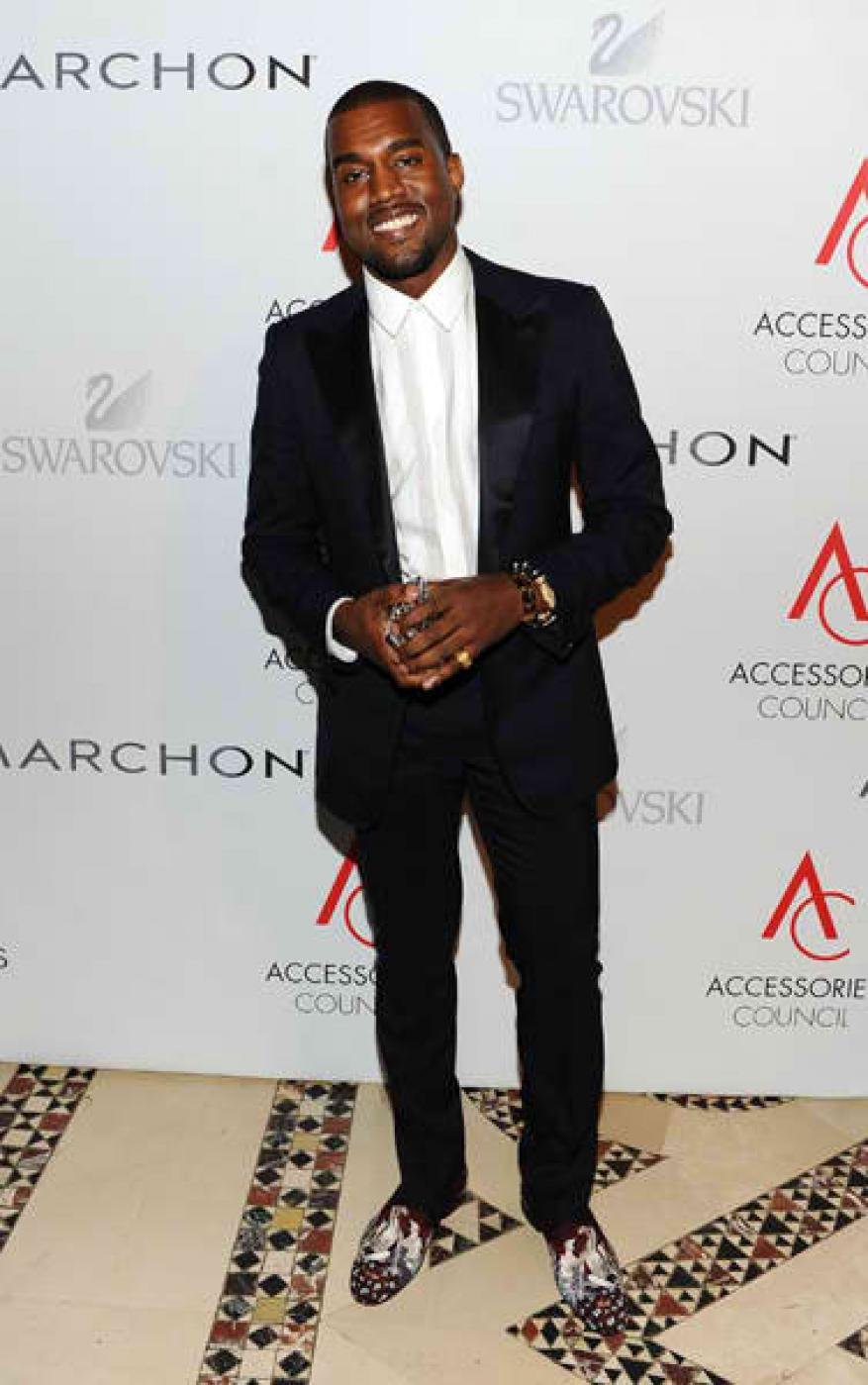 Kanye West smiling in a dark suit, white button-down shirt, and patterned loafers standing against a branded step-and-repeat backdrop
