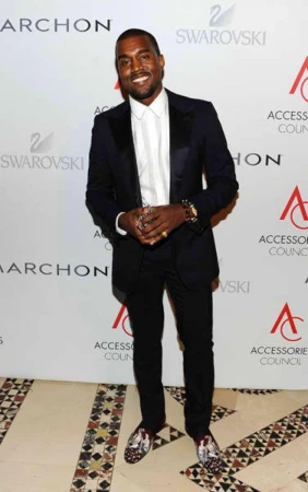 Kanye West smiling in a dark suit, white button-down shirt, and patterned loafers standing against a branded step-and-repeat backdrop