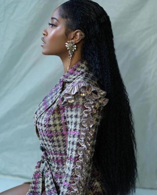 Profile shot of a Black woman with long hair wearing a houndstooth blazer with ruffles, and statement earrings, against a gray backdrop.