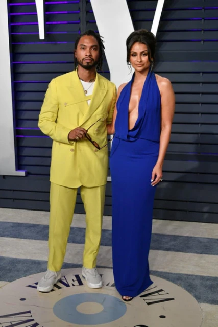 Full shot of a stylish couple in formal wear posing against a purple backdrop at an event. A man in a yellow suit stands next to a woman.