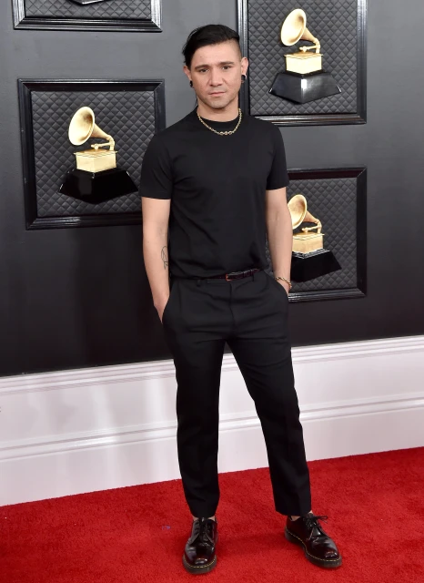 Full-length shot of a fair-skinned adult male with dark hair, wearing a black shirt and pants, standing on a red carpet. 