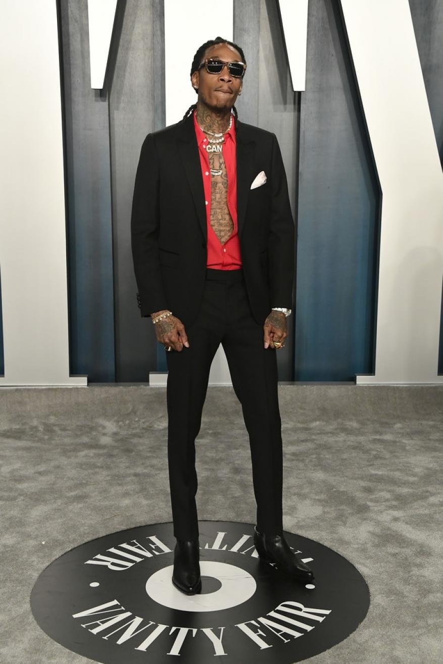 Wiz Khalifa poses in a black suit with a red shirt and patterned tie on a Vanity Fair backdrop at an indoor event, full shot.