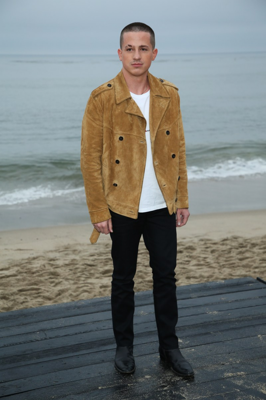 Charlie Puth wearing a camel suede jacket and black trousers standing on a wooden platform at the beach.