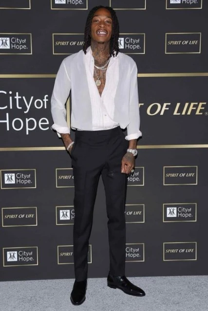 Full body shot of Wiz Khalifa on red carpet wearing a white blouse, black pants and dress shoes, wrist jewelry, necklaces, with tattoos.