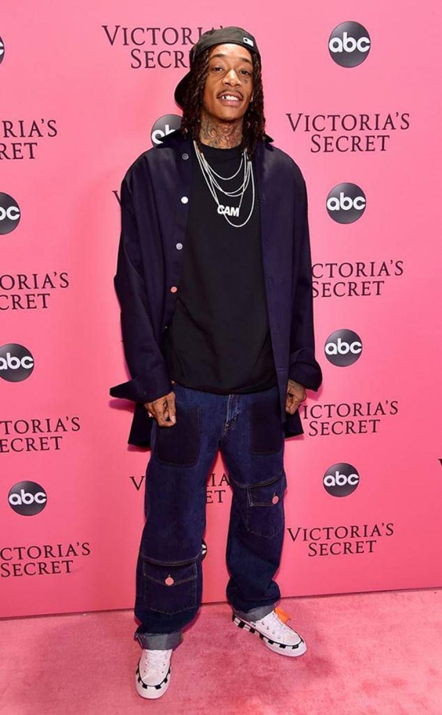 Adult male wearing a baseball cap, black t-shirt, denim jacket, dark jeans, and white sneakers posing in front of a Victoria's Secret backdrop.