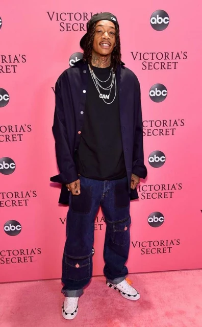 Adult male wearing a baseball cap, black t-shirt, denim jacket, dark jeans, and white sneakers posing in front of a Victoria's Secret backdrop.